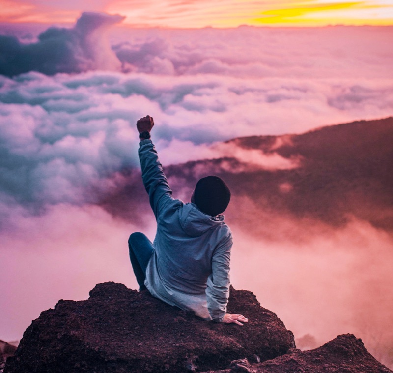 Photo d'un homme de dos, assis au sommet d'une montagne qu'il a gravi. Il lève le poing par fierté. Illustration utilisée dans le cadre d’une formation communication positive proposé par Audrey Martin Coaching. Disponible en présentiel dans les Landes et la Gironde, ou à distance.